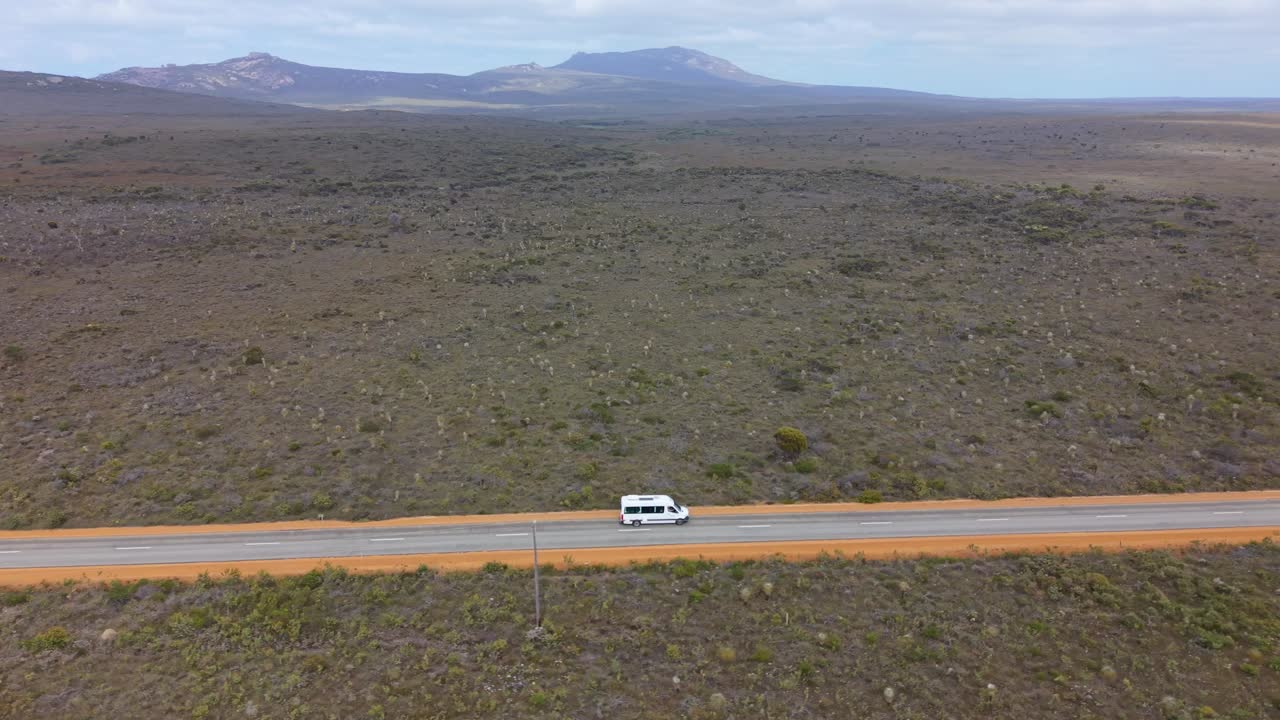 Vista aérea de drone de um motorhome dirigindo por uma rodovia solitária em uma paisagem semidesértica com colinas distantes.