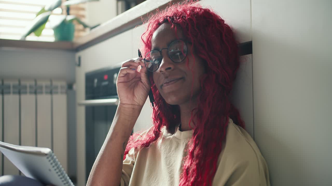 Ragazza afroamericana con capelli ricci rossi che studia sul pavimento della cucina, prendendo appunti, guardando la fotocamera e sorridendo