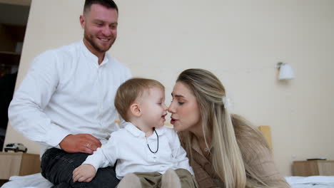 adorable baby boy sitting on the bed looks at his mommy. mother and son touch noses lovingly. close up. bearded smiling man sits at backdrop. low angle view.