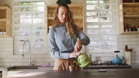 Happy-caucasian-woman-standing-at-counter-in-cottage-kitchen-pouring-tea-from-teapot-and-smiling