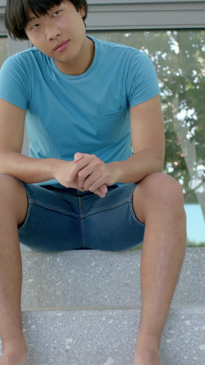Vertical video: Asian male teen sitting on concrete steps resting, shifting gaze at glass panels
