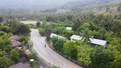 Aerial-drone-of-motorbike-on-rough-gravel-road-surrounded-by-green-trees-on-rural-tropical-Atauro-Island,-Timor-Leste,-South-East-Asia