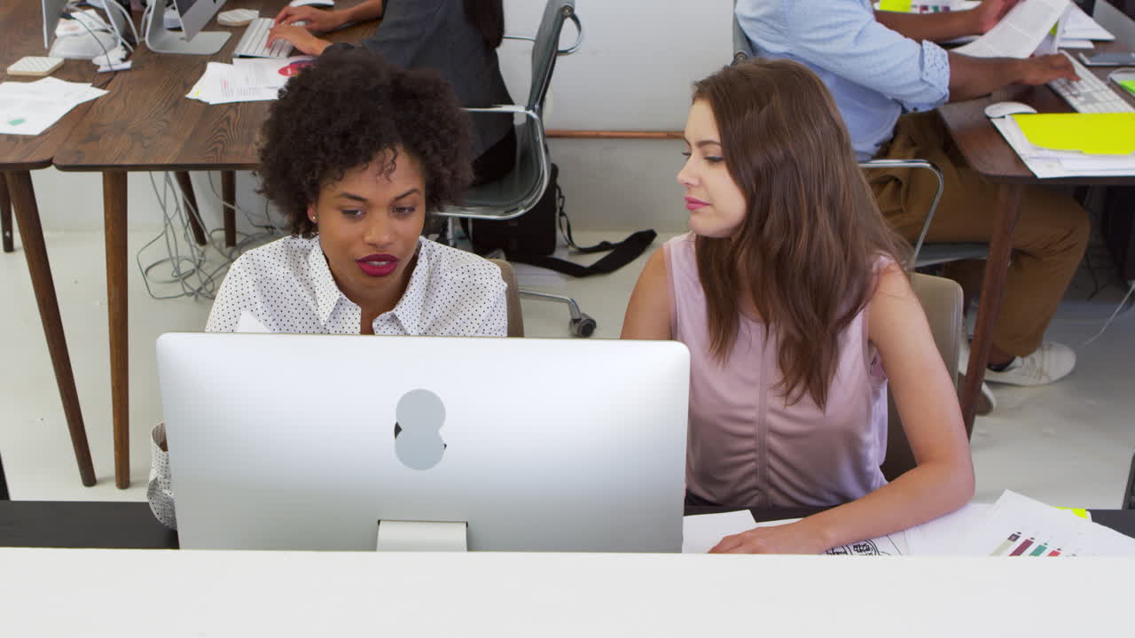 Two happy women discuss work at computer in open plan office, elevated view