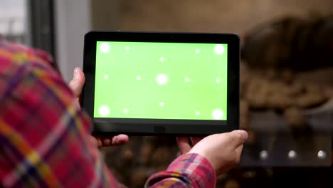 primer plano, el agricultor sostiene una tableta digital con pantalla verde en sus manos, en el fondo del almacén de almacenamiento de patatas. agricultura, industria alimentaria, cosecha