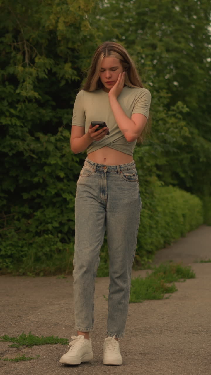Woman in crop top, revealing her midriff, walking along greenery-lined path while focused on her phone, surrounded by trees and a wooden bench in background, with a distant building visible