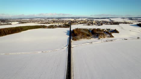 Vista-Aérea-De-Una-Carretera-Recta-Que-Conduce-A-La-Ciudad-Mientras-Los-Coches-Pasan-Por-El-Paisaje-Rural-Nevado