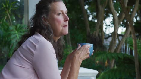 Thoughtful-senior-caucasian-woman-standing-on-terrace-drinking-coffee-and-looking-around