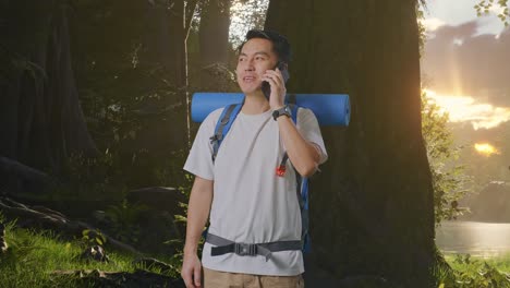 asian male hiker with mountaineering backpack smiling and talking on smartphone while exploring forest nature