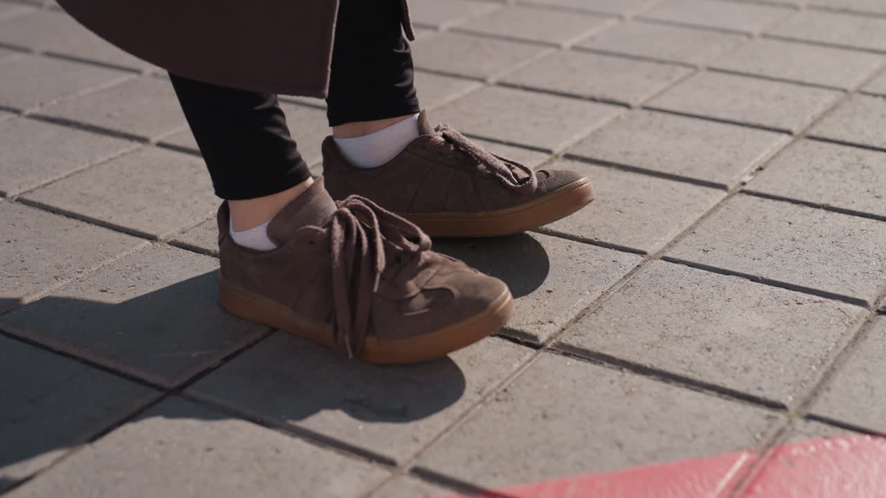 Brown sneakers crossing red painted line on tiled pavement, closeup of ankles and shadows, casual commuter stride, textured concrete tiles bathed in warm sunlight, minimal urban composition