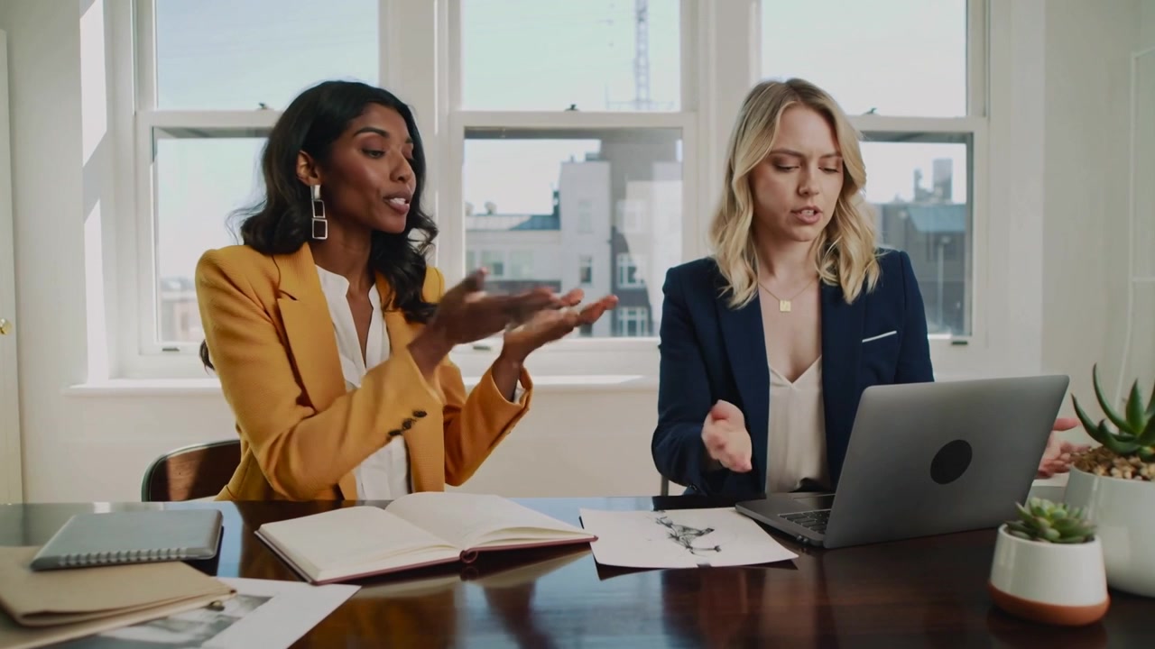 Mentre sfoglia un taccuino, una donna in blazer inizia una revisione in ufficio mentre un collega risponde al portatile.