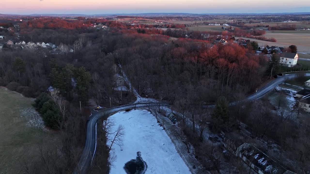 American suburban landscape at sunset, showing frosty fields, leafless trees and scattered homes. Warm evening light contrasts with cold rural winter scenery. Aerial flyover wide shot