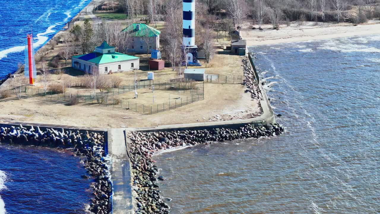 A black and white lighthouse rises beside simple houses on a long stone jetty splitting vivid blue Baltic Sea from muted river waters, bordered by sand and leafless trees.