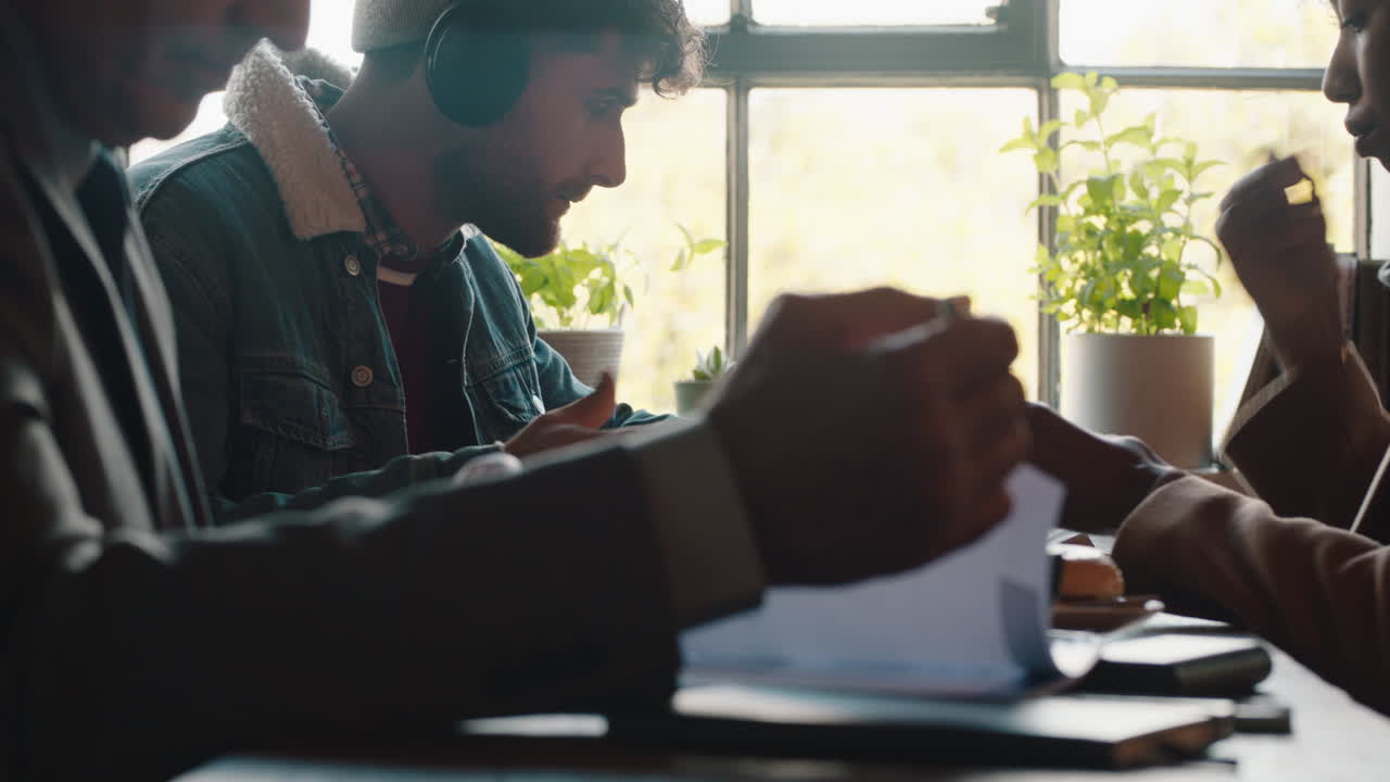 young hipster man using smartphone drinking coffee in cafe texting sharing messages on social media wearing headphones enjoying listening to music in busy restaurant