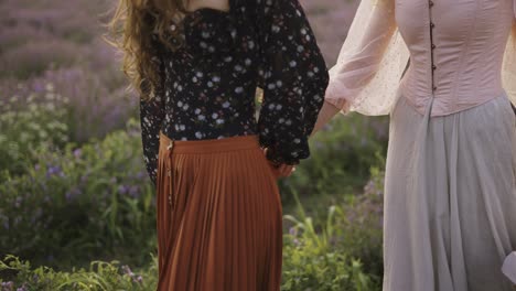 Two-curly-women-travelers-are-walking-along-a-lavender-field,-holding-hands