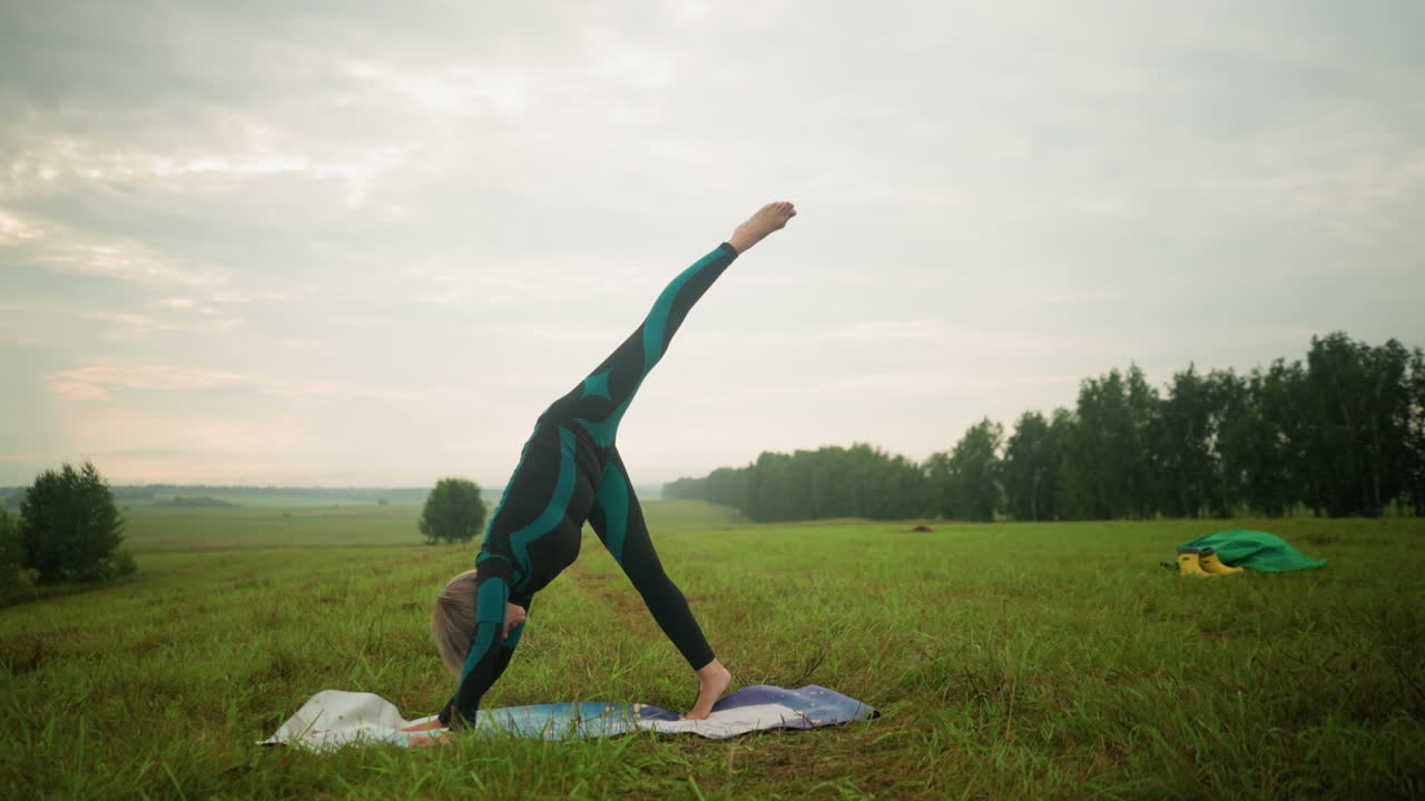 mujer en traje verde y negro practicando postura de yoga dividida de pie al aire libre en una alfombra de yoga en un campo cubierto de hierba bajo un cielo nublado, estirando su cuerpo hacia abajo como parte de una rutina de bienestar