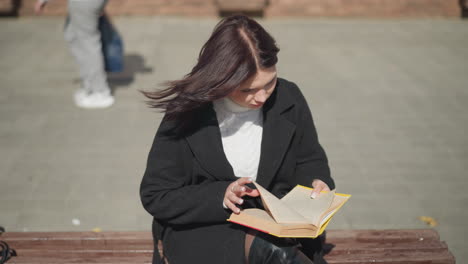 woman in black coat sitting outdoors, engrossed in her book, she flips to a new page while her hair flutters in the wind, a passerby is seen in the background from behind