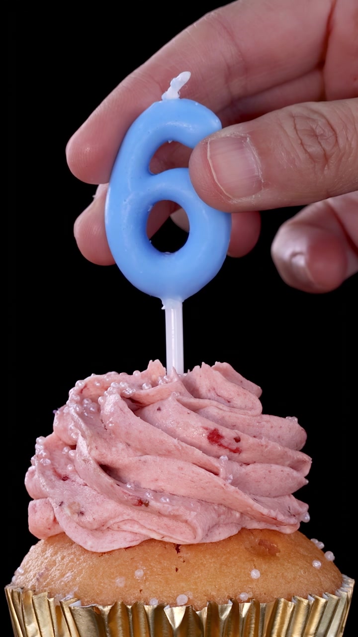 Una mano inserta una vela de cumpleaños azul con el número seis en un cupcake de fresa glaseado con envoltorio dorado, bajo una brillante iluminación de estudio y fondo negro.
