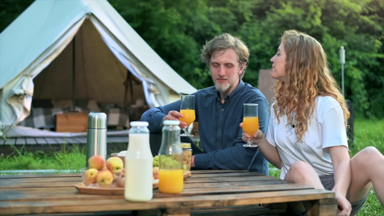 Una coppia che fa colazione immersa nella natura in un glamping. Bevande, cibo, chiacchiere e sorrisi. Tenda sullo sfondo. Movimento al rallentatore
