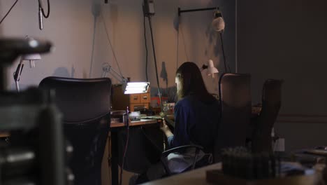 back view of caucasian female jeweller sitting at desk, making jewelry in workshop