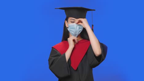 retrato de una estudiante en traje de graduación con gorra y túnica pone una máscara de protección médica. joven mujer morena posando en el estudio con fondo de pantalla azul. primer plano. cámara lenta lista 59.94fps