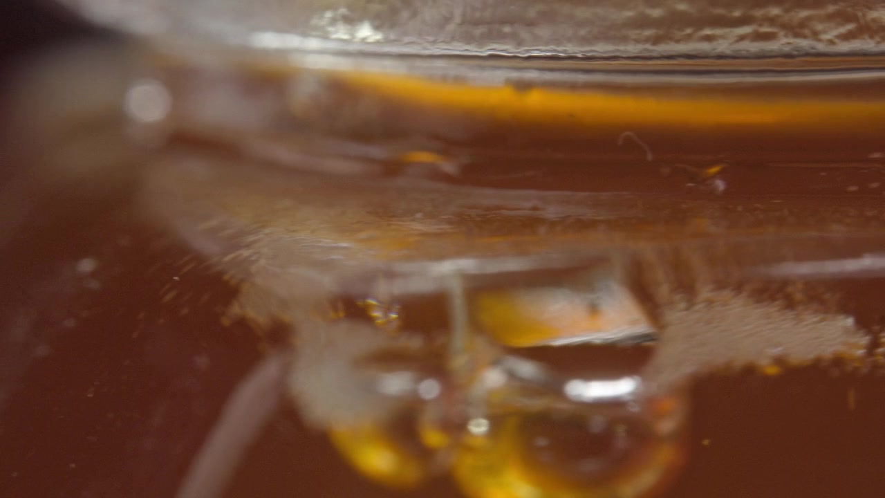 Una panoramica si muove attraverso un barattolo di vetro di miele riempito di miele dorato, rivelando il suo ricco colore ambrato, la consistenza lucida, i riflessi e la densa consistenza liquida in vista macro.