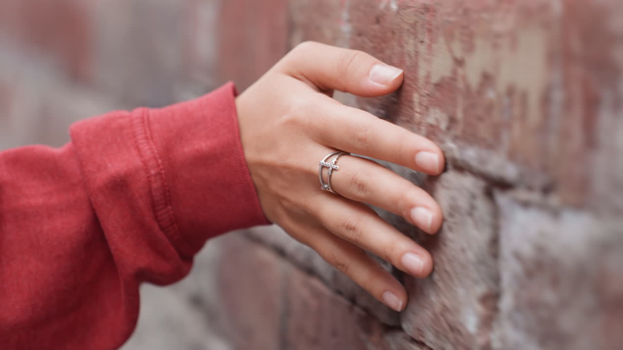 Mano di donna nera che tocca mattone stagionato, primo piano delle dita con anelli d'argento, manica di maglione rosso nell'inquadratura, malta testurizzata e sottile patina, gesto lento e intimo che si concentra su gioielli e superficie