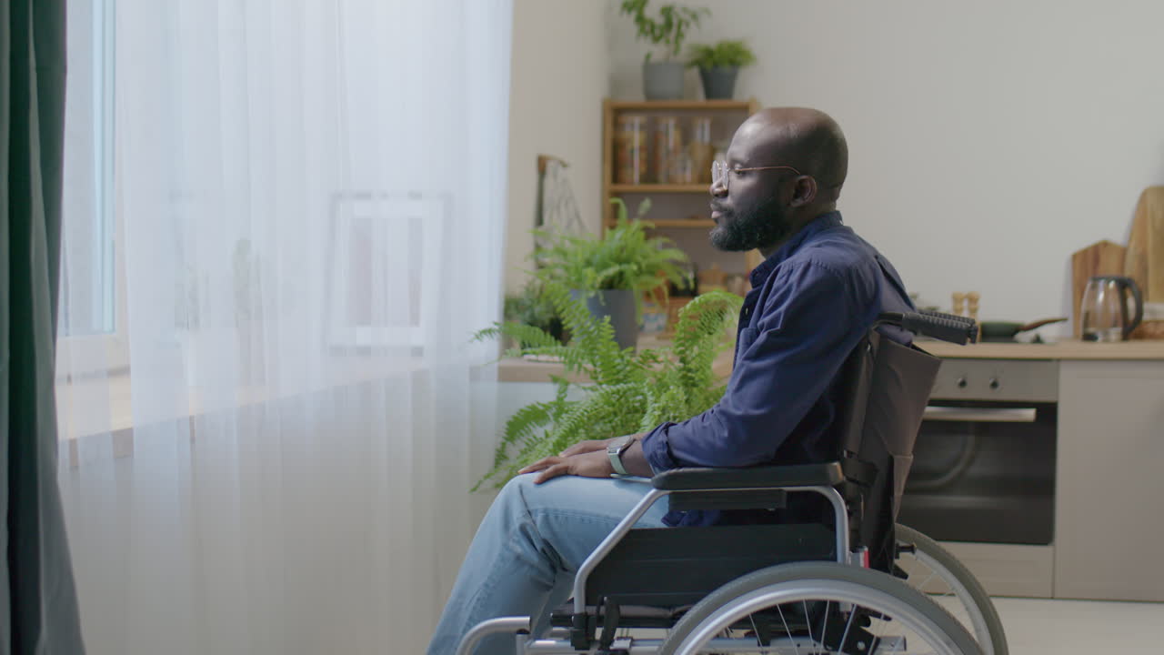 Man in Wheelchair Rubbing Eyes and Looking through Window at Home