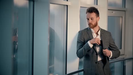 Un-Joven-Elegante-Parado-Junto-A-La-Ventana-En-El-Fondo-De-La-Ciudad-Nocturna-Recibe-Un-Mensaje-Alegre-En-Un-Smartphone