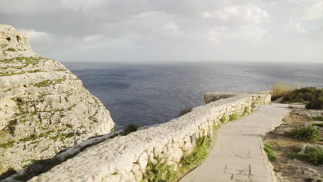 Scenic-seaside-stone-footpath-and-rock-wall-with-picturesque-view-of-mountain-face-and-blue-ocean-sea-water-and-sky,-Malta,-handheld-pan-forward