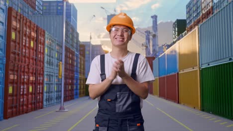 un trabajador asiático con gafas de protección y casco de seguridad sonriendo y aplaudiendo sus manos mientras está de pie en el almacén del patio de contenedores