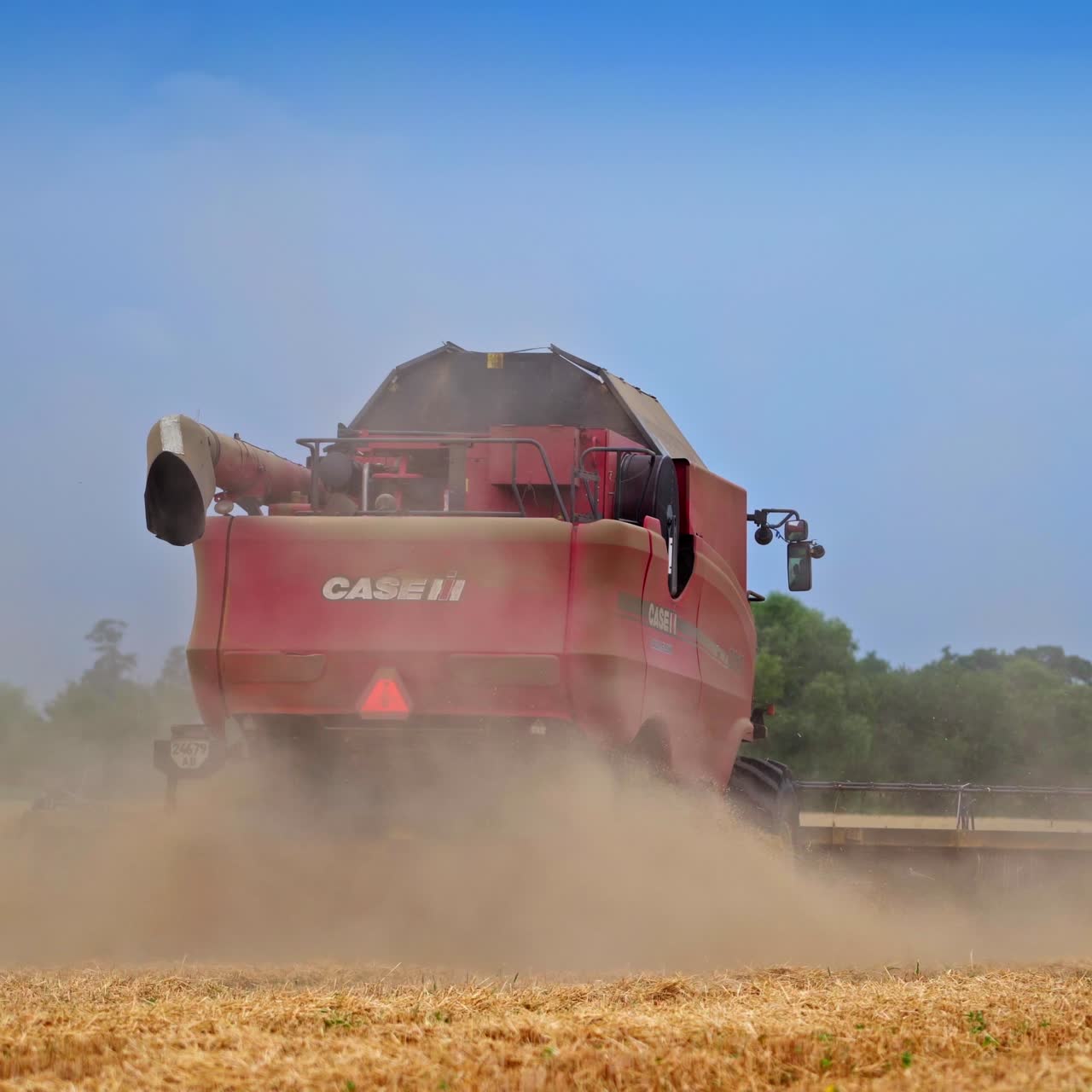 The back of the big red harvesting machine. Combine working and making the clouds of dust. Green trees and blue sky backdrop