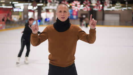 middle-aged bald man in a brown sweater and black scarf joyfully dancing on an ice rink, pointing upwards, with blurred skaters in the background