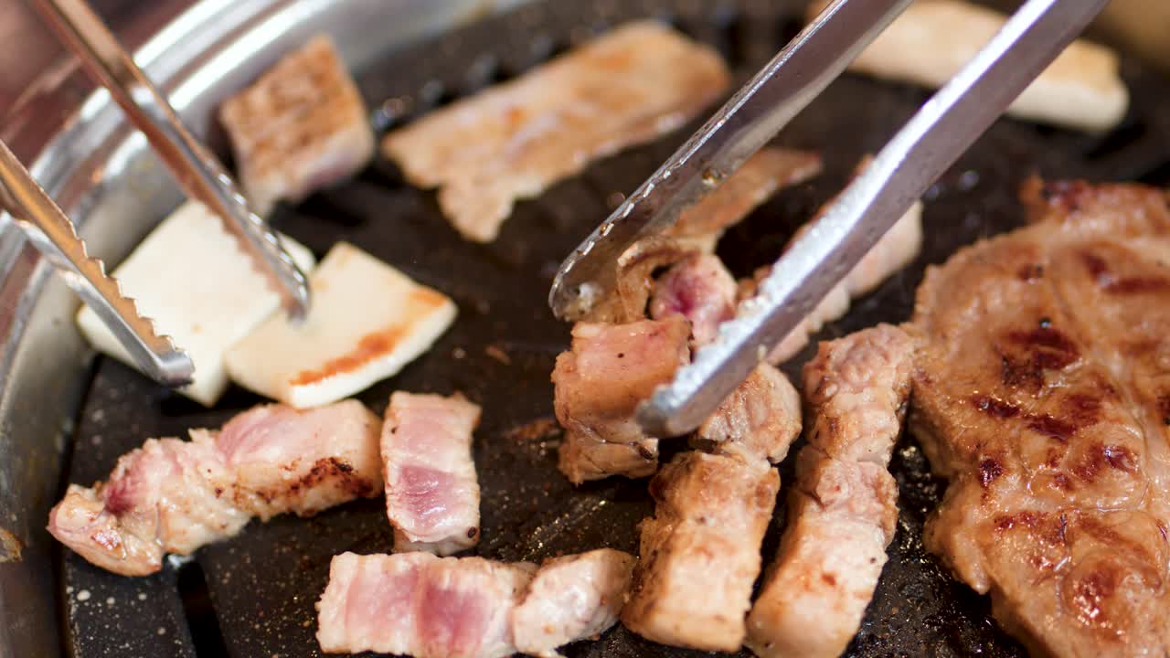 Tongs turn sizzling pork belly and cheese on a hot Korean barbecue grill, close-up view