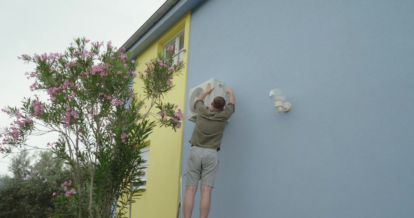 Un homme réparant une unité de climatisation extérieure montée sur le mur d'une maison. L'accent est mis sur les travaux d'entretien, le service technique et la réparation de systèmes de refroidissement domestiques.