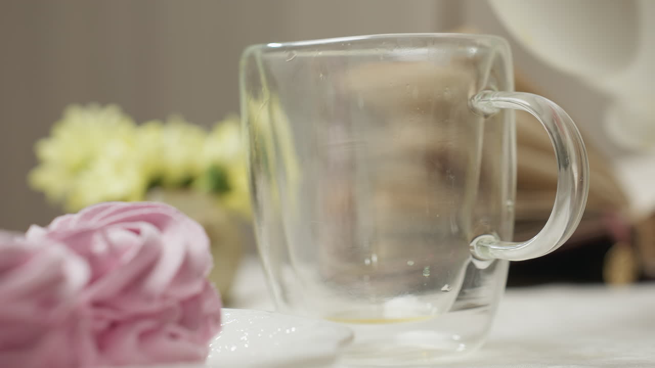 Close up glass cup with drops of herbal tea as person pours from white ceramic teapot beside soft pink cupcake on white plate with small yellow flowers