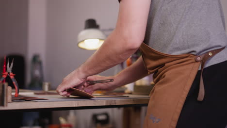 Tailor-with-apron-cuts-rectangle-detail-of-leather-at-desk