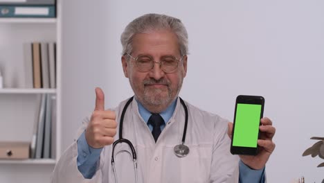 un médico de edad avanzada sostiene un teléfono inteligente con una tecla de croma de pantalla verde, lo señala con el dedo y hace un gesto con el pulgar hacia arriba. un médico con una bata médica blanca se sienta en la mesa en la oficina del hospital.