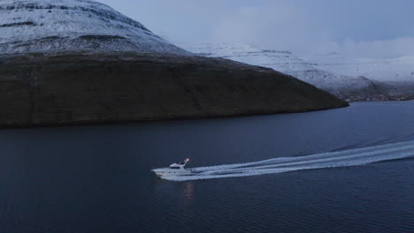 faroe islands, 4k aerial tracking pan of fishing boat with snow covered mountains in the background as it dissapears into the sun