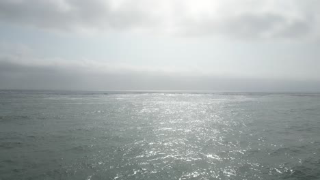 low angle aerial drone shot over the sea on a sunny day reflecting the sun on the water with view of the horizon with surfern with surfboards in water