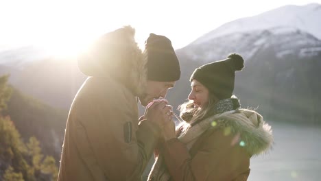 Young-couple-in-winter-coats-standingto-each-other,-holding-hands-towards-gorgeous-snowy-mountains-and-lake,-a-man-warming-his-girlfiends-hands-and-kissing.-Travel-concept.-Sun-strongly-shining-on-the-background