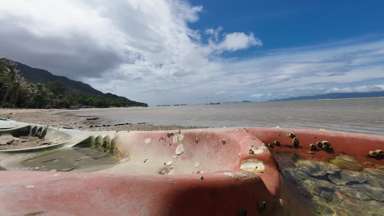 Timelapse footage of an old stranded kayak covered in seashells and algae on a sandy beach of Koh Phangan Thailand with moving clouds and scenic tropical seascape background