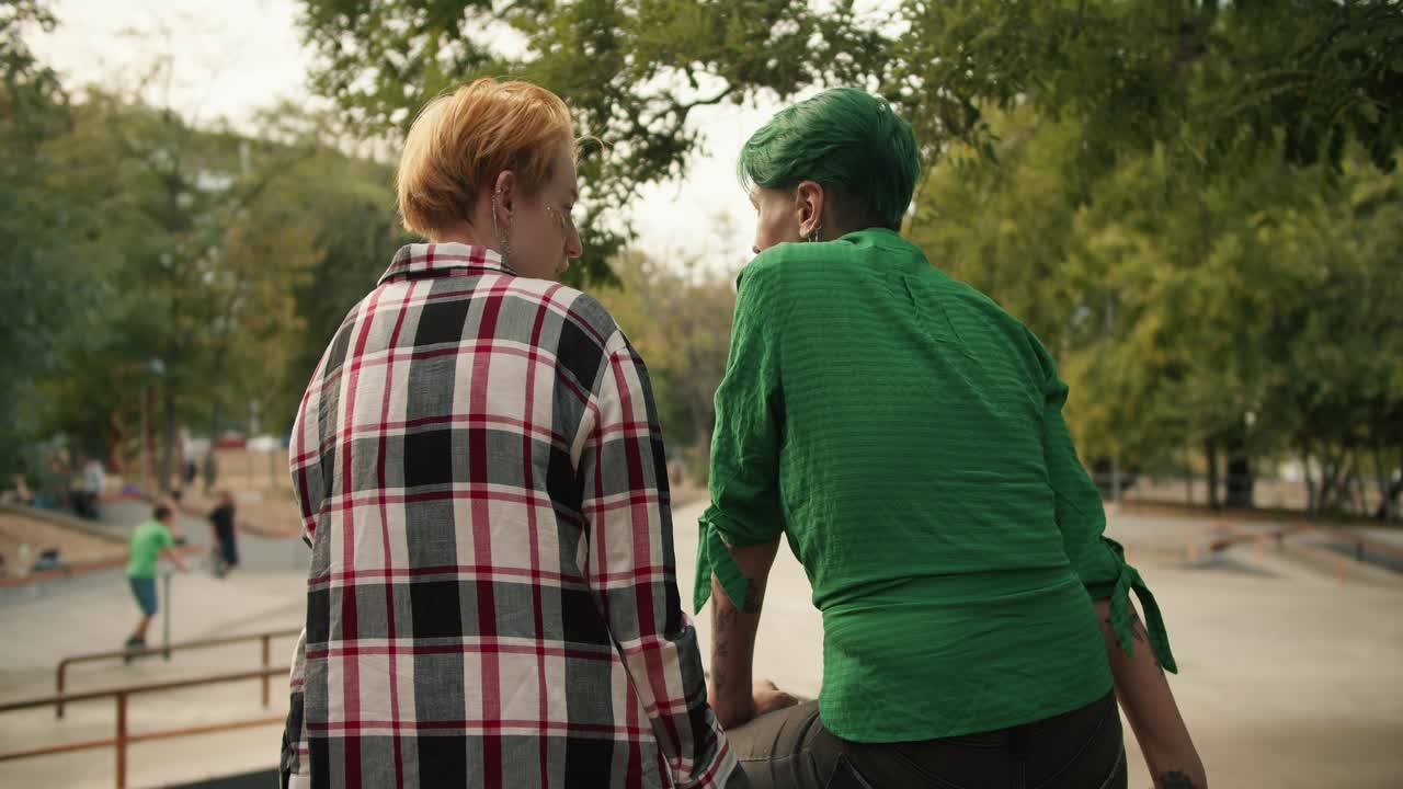 Rear view of a girl with dark yellow short hair in a plaid shirt and a girl with short green hair in a green shirt sitting on the fence near the skatepark and talking in the park in the summer