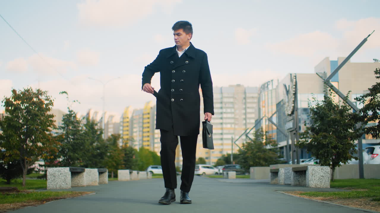 Teacher in black coat walking confidently outdoors, reaching into pocket to retrieve phone and answer call, surrounded by urban greenery and blurred modern buildings under bright daylight