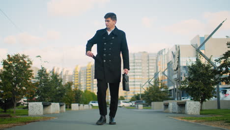 teacher in black coat walking confidently outdoors, reaching into pocket to retrieve phone and answer call, surrounded by urban greenery and blurred modern buildings under bright daylight