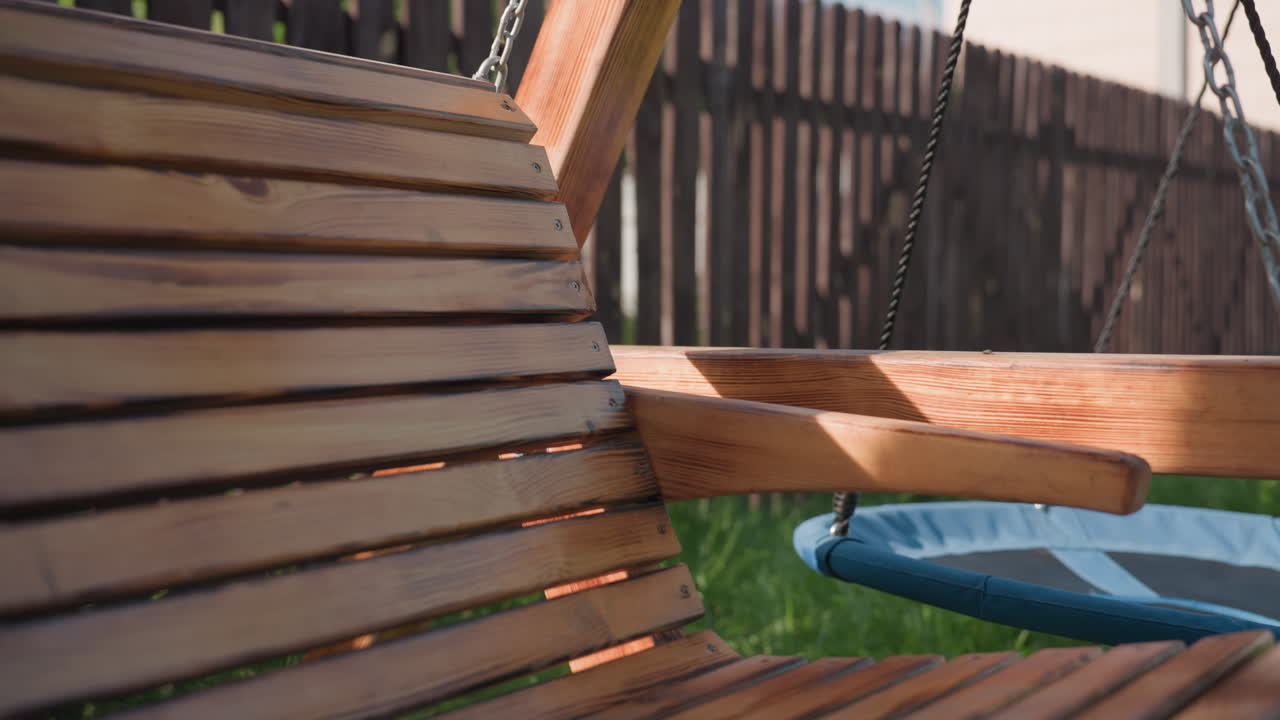 Close up of smooth wooden swing suspended by chain gently moving beside second swing, sunlight casting soft shadows across warm wood tones with dark fence creating contrast in quiet backyard