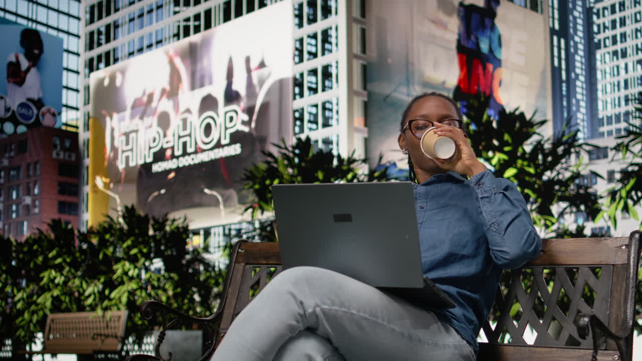Remote worker drinking coffee, using laptop on bench in the park