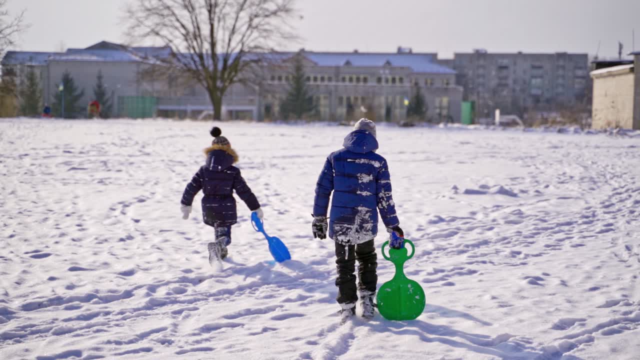 Frost winter season. Two children walk a winter road with plastic spade sledges. Winter games and fun. Kids having winter vacation. Slow motion.