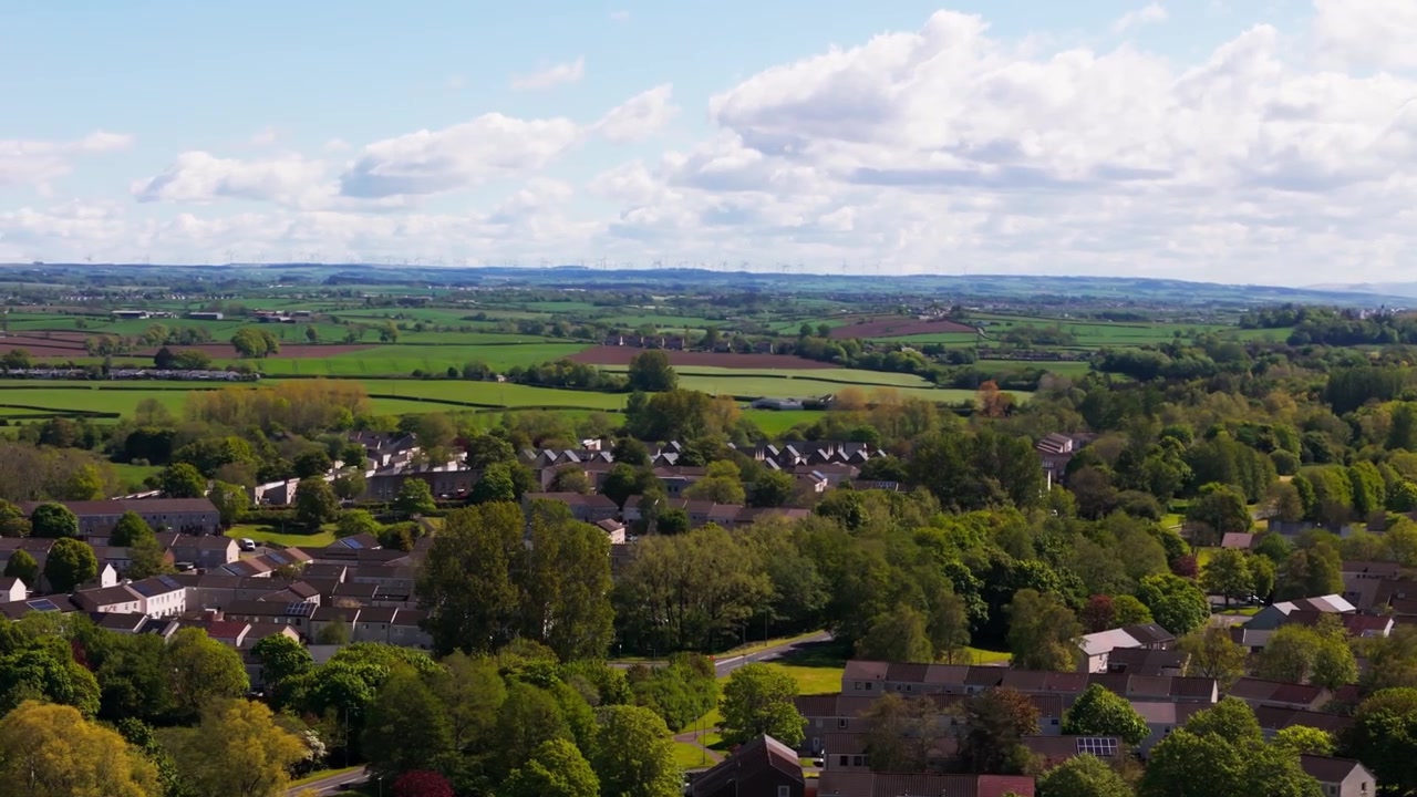 Countryside village surrounded by farmland and rolling green landscape in Scotland under bright daylight and scattered cloud, cinematic aerial view
