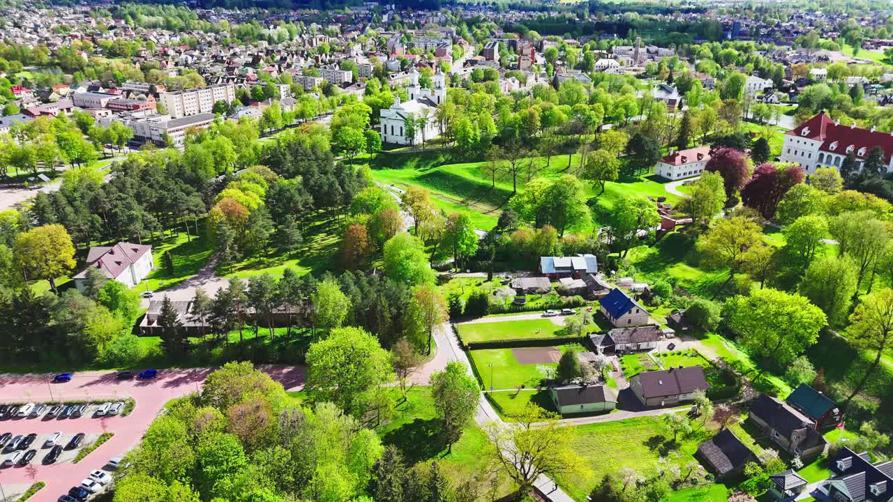 Vibrant green countryside with scattered houses transitions into a more densely packed city in the background, offering a contrast of rural and urban life from above.