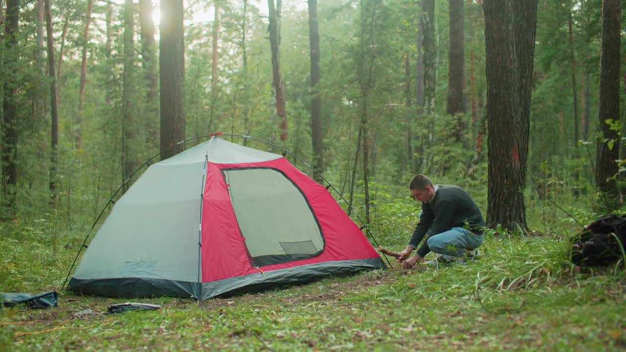 man crouches near red and gray tent in dense green forest using thick wood to hammer peg into ground while setting up campsite during daylight with trees and scattered camping gear around